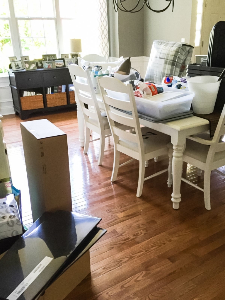 A dining room with white chairs and a table covered in plastic bins, boxes, and assorted items. Sunlight streams in through large windows. Unpacked boxes are on the wooden floor.