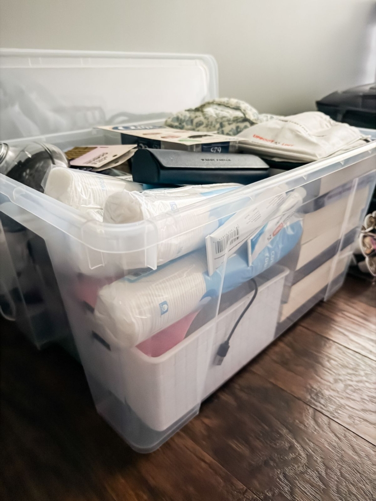 A clear plastic storage bin filled with various household items, including paper cups, books, boxes, and towels, sits on a dark wooden floor with its lid resting behind it.