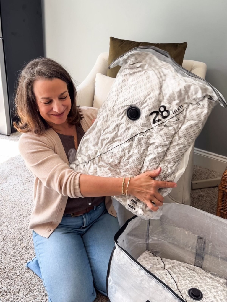 A woman kneels on carpet, smiling as she holds a large vacuum-sealed storage bag labeled &ldquo;28,&rdquo; next to an open storage container.