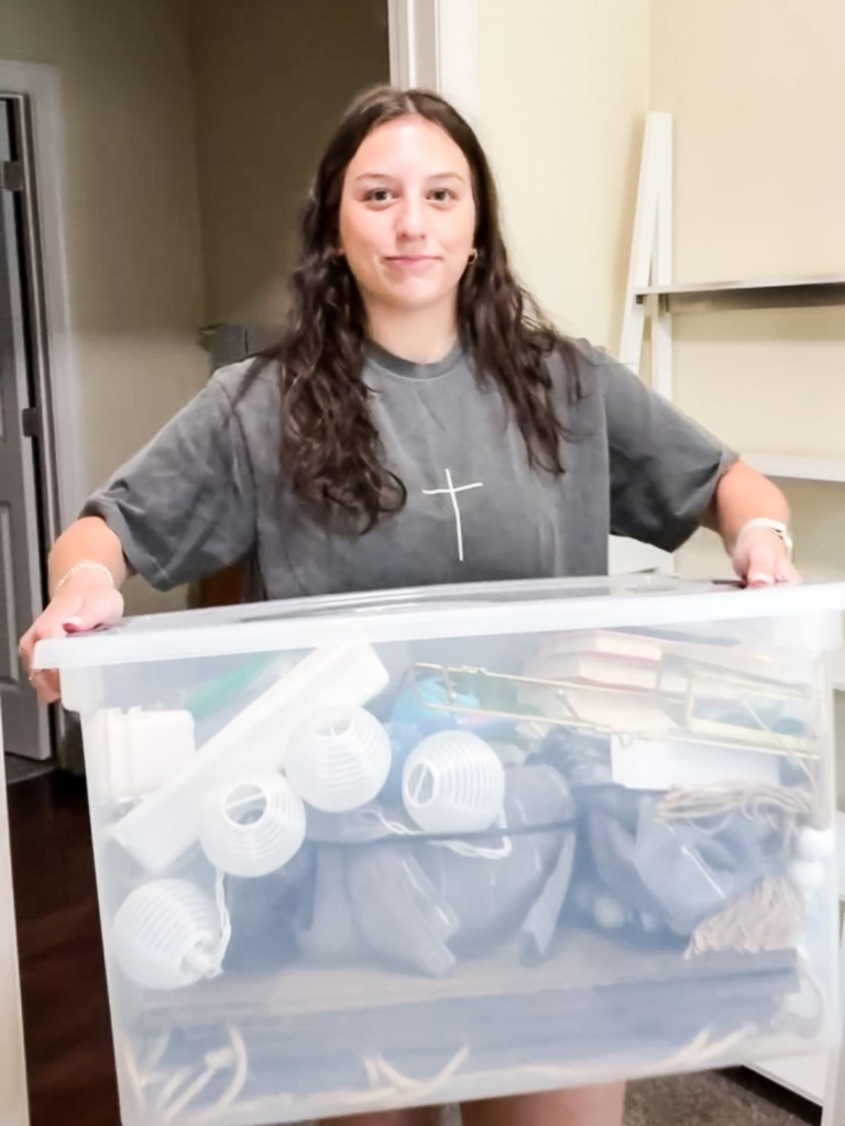 A young woman with long brown hair holds a large clear plastic bin filled with various household items, including white lanterns and books, standing indoors near an open doorway.