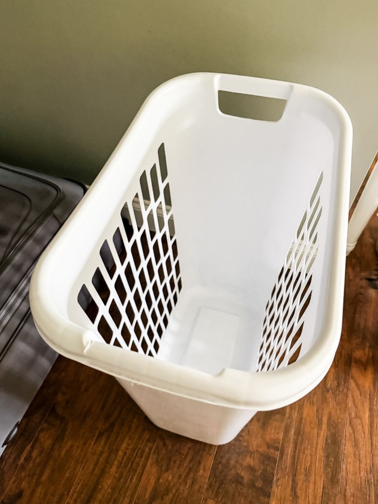 A white plastic laundry basket with holes on the sides sits empty on a wooden floor next to a green wall.