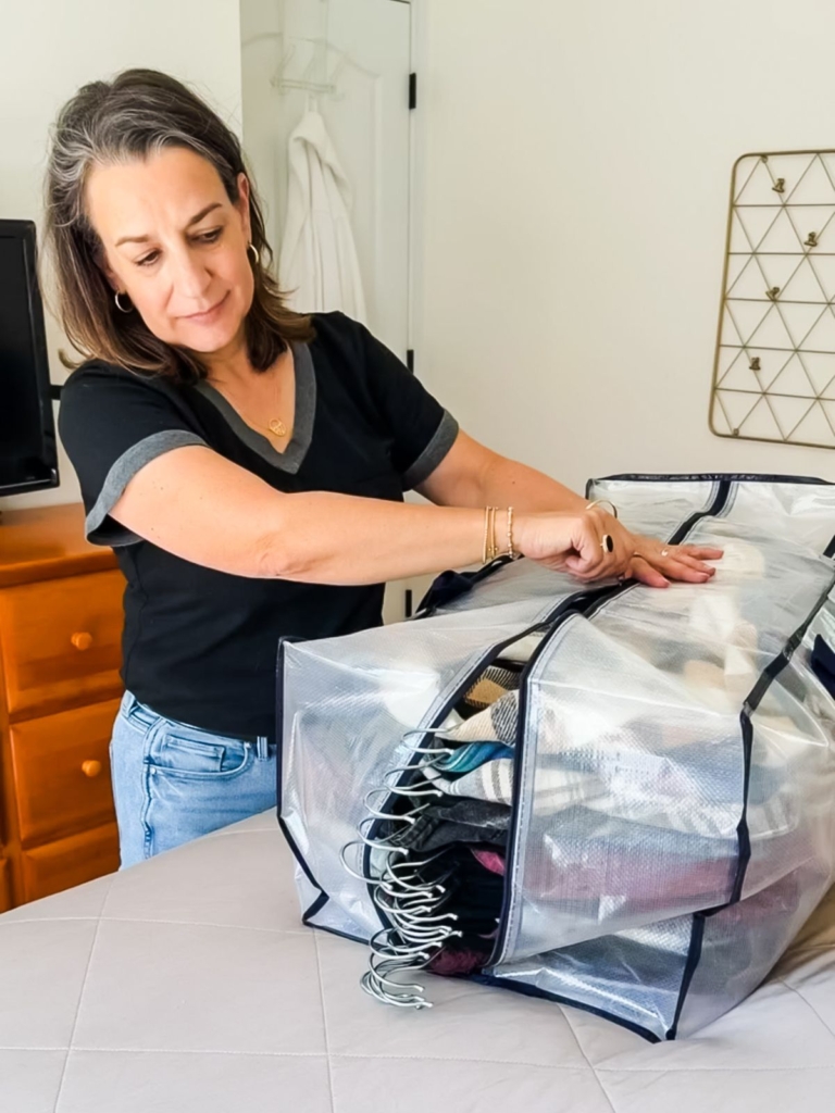 A woman in a black shirt zips up a large, clear storage bag filled with folded clothes on a bed in a bright bedroom.