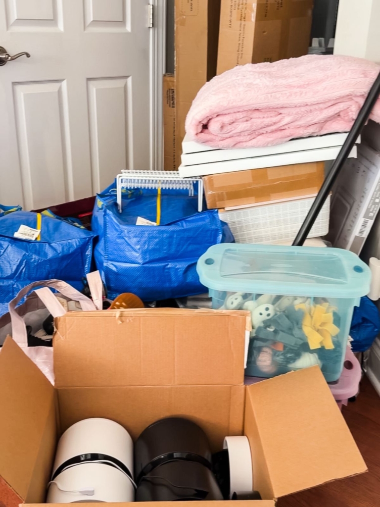Stacked blue bags, a cardboard box with kitchen items, a pink blanket, and a clear bin of toys are arranged near a door with several cardboard boxes. The scene suggests packing or moving.