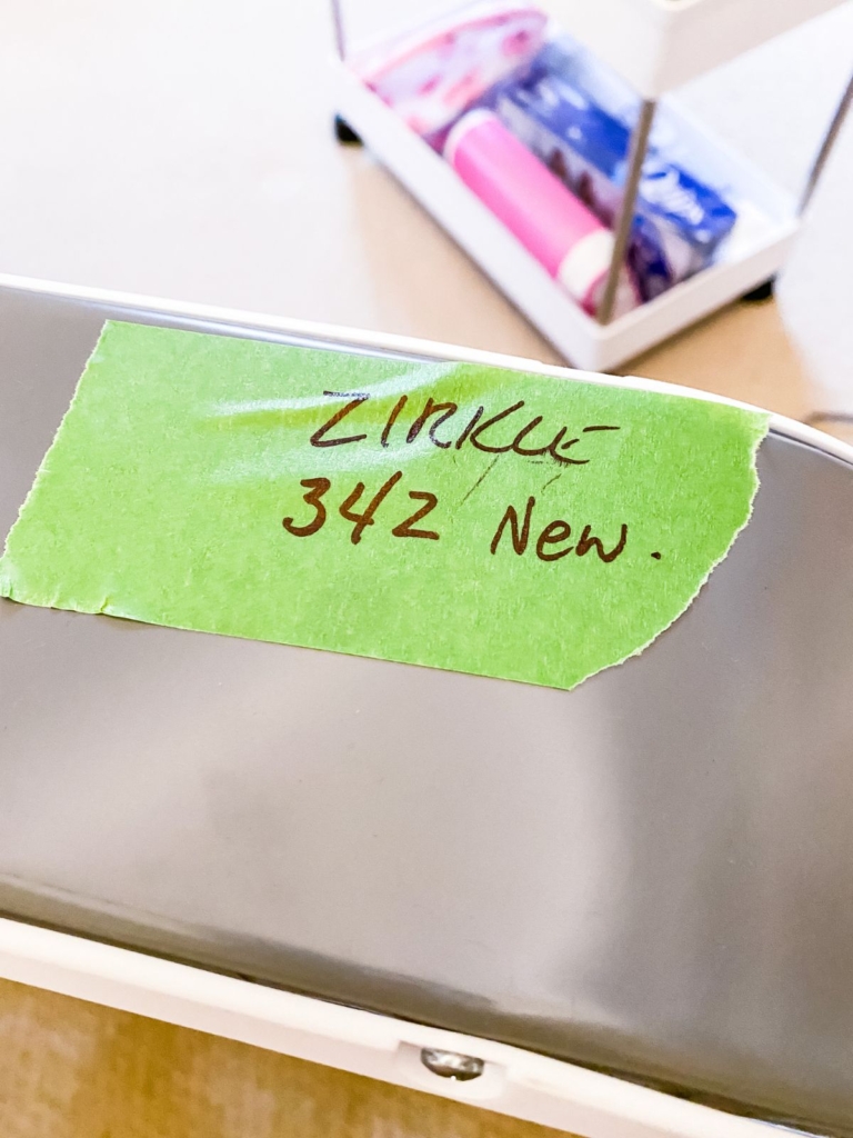 A piece of green tape labeled ZIRKLE 342 New. is stuck to the edge of a gray surface. In the background, a cart with various items is slightly out of focus.