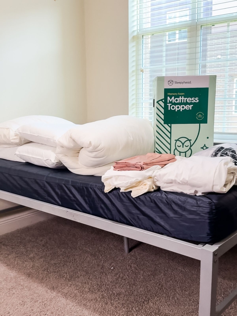 A mattress topper box, folded bedding, and linens are placed on a bare mattress of a metal bed frame in a bright room with carpeted floors and a window.