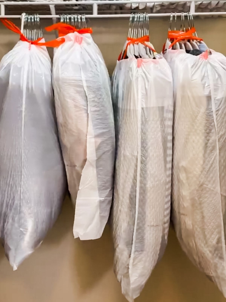 Four large plastic trash bags filled with hanging clothes are tied with orange ties and hung on a metal closet rod.