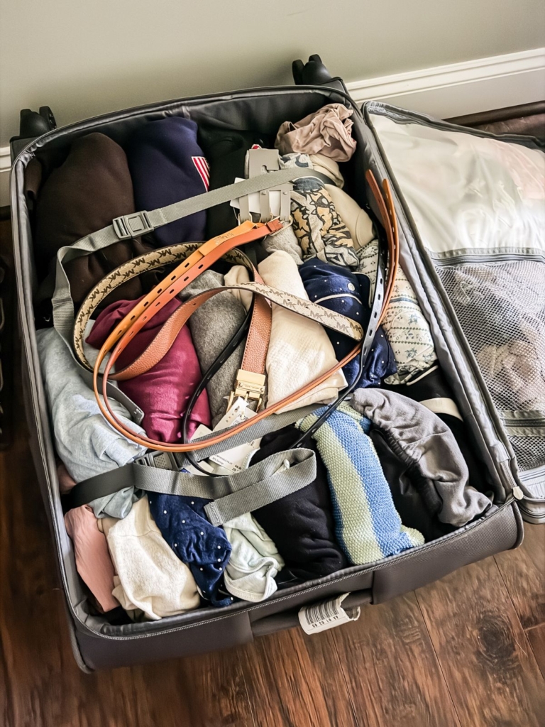 An open suitcase filled with neatly packed clothes, several belts laid across the top, and a mesh compartment on the lid. The suitcase is on a wooden floor next to a wall.