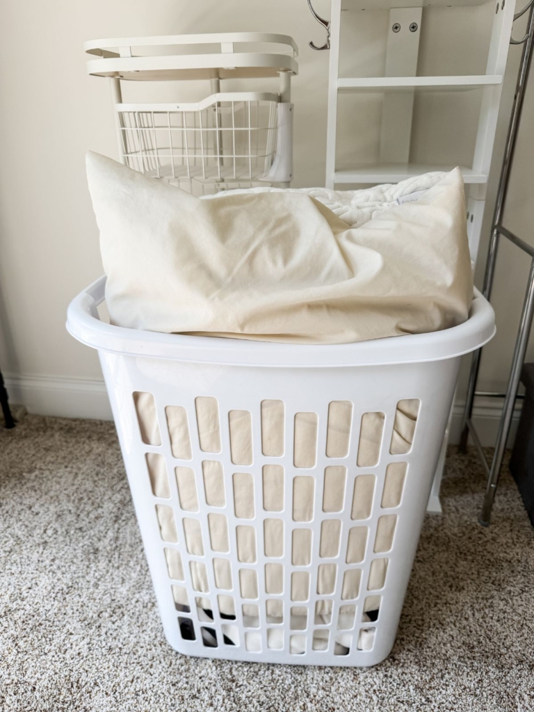 A white laundry basket filled with beige bed linens sits on a carpeted floor, with shelves and a laundry cart in the background.
