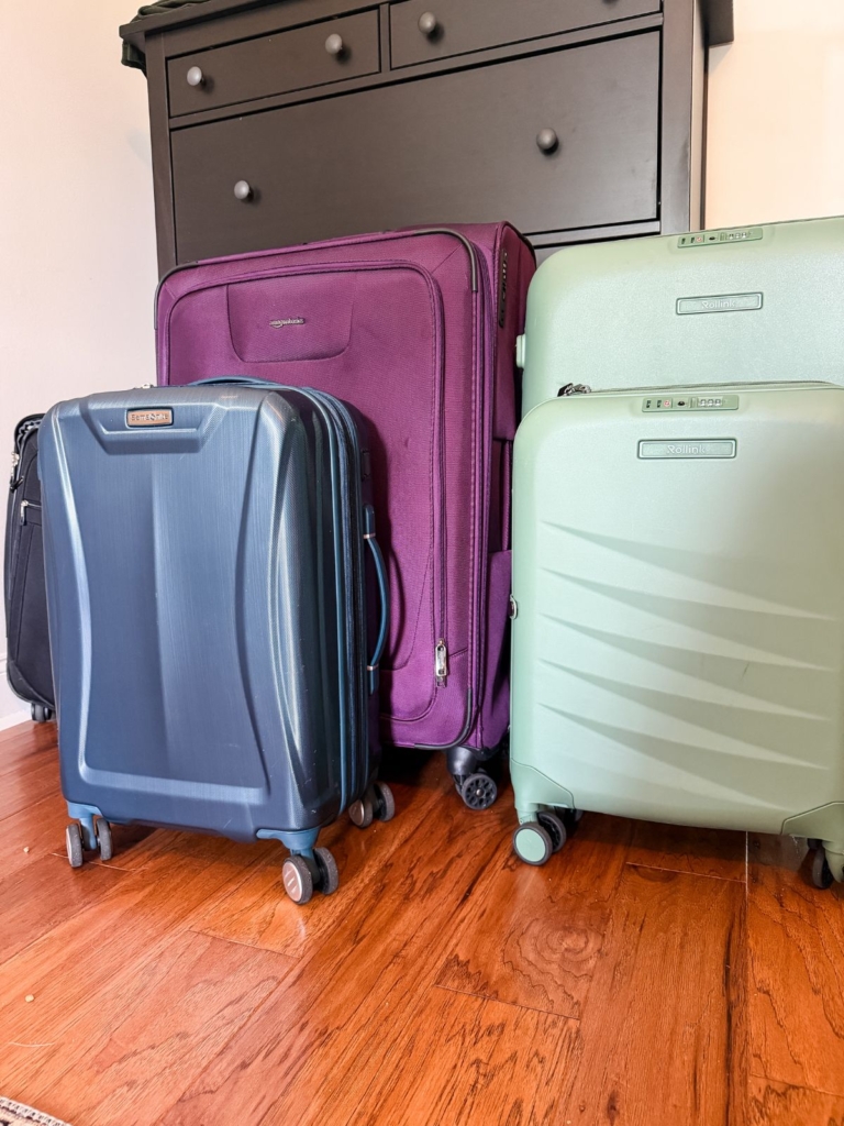 Four suitcases&mdash;one blue, one purple, and two light green&mdash;stand upright on a wooden floor in front of a black dresser. The suitcases are various sizes and have wheels.