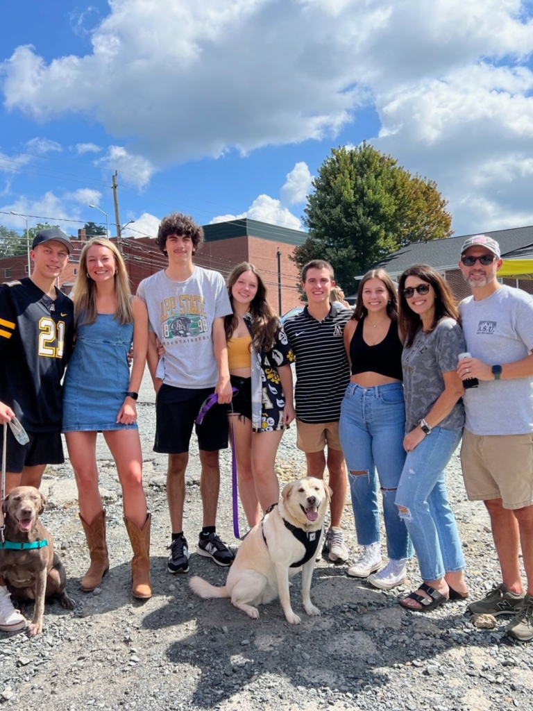 A group of seven people and two dogs pose outdoors on a sunny day. The group is smiling, dressed casually, and standing on gravel with trees, buildings, and blue sky in the background.