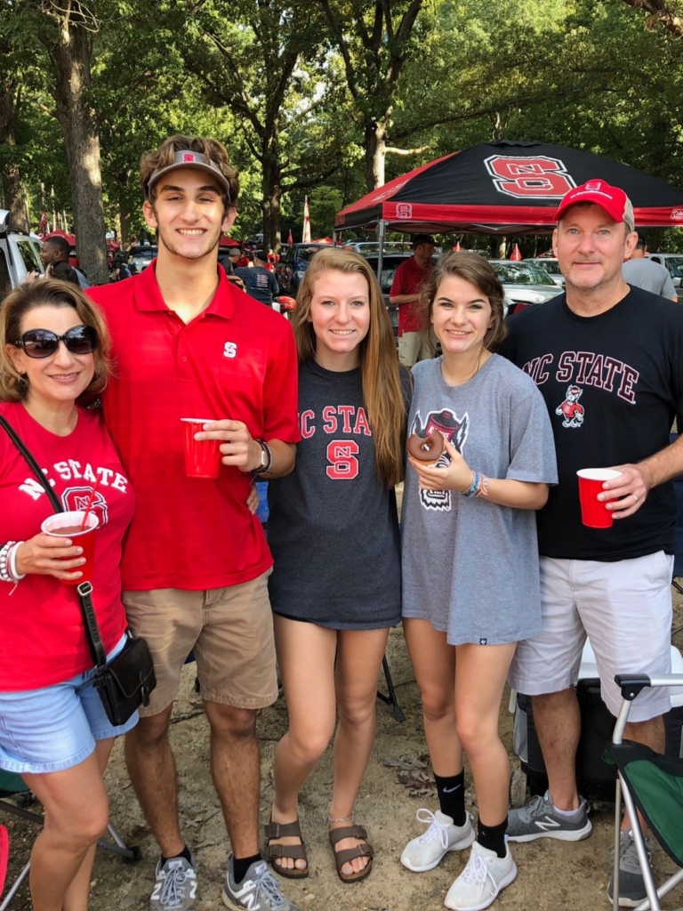 Five people wearing NC State gear smile at a tailgate. They stand outdoors near tents and trees, holding drinks and posing together, showing school spirit and camaraderie.