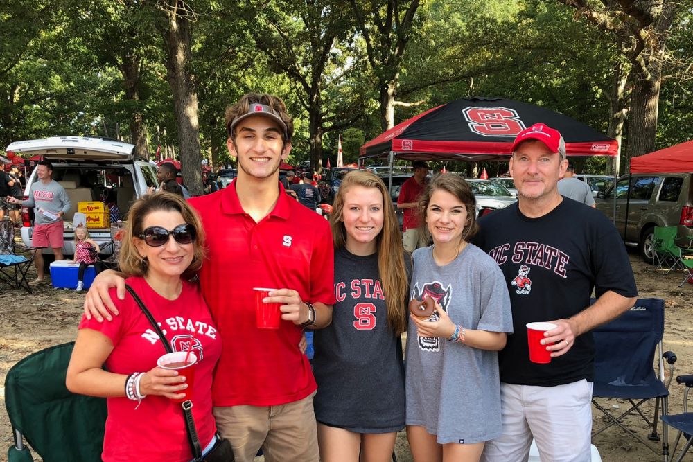 Five people wearing NC State attire smile at an outdoor tailgate, standing in front of tents and parked cars, holding red cups. Trees and other tailgaters are visible in the background.