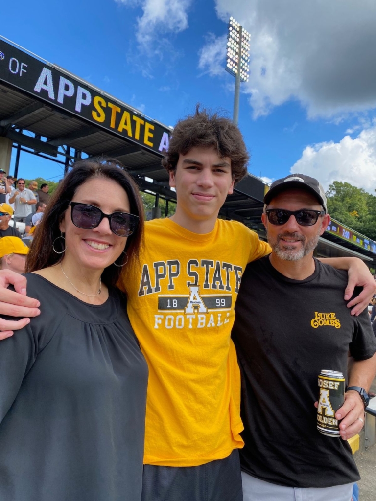 Three people, two men and one woman, stand arm-in-arm smiling at an outdoor stadium. They wear sunglasses and App State apparel, with a crowd and a sign reading APP STATE visible in the background.