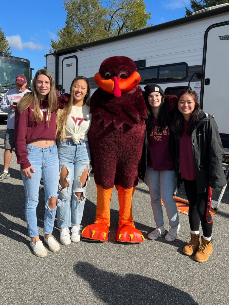 Four young women stand smiling with the Virginia Tech HokieBird mascot outside, wearing casual clothes and VT gear, with a trailer and trees in the background.