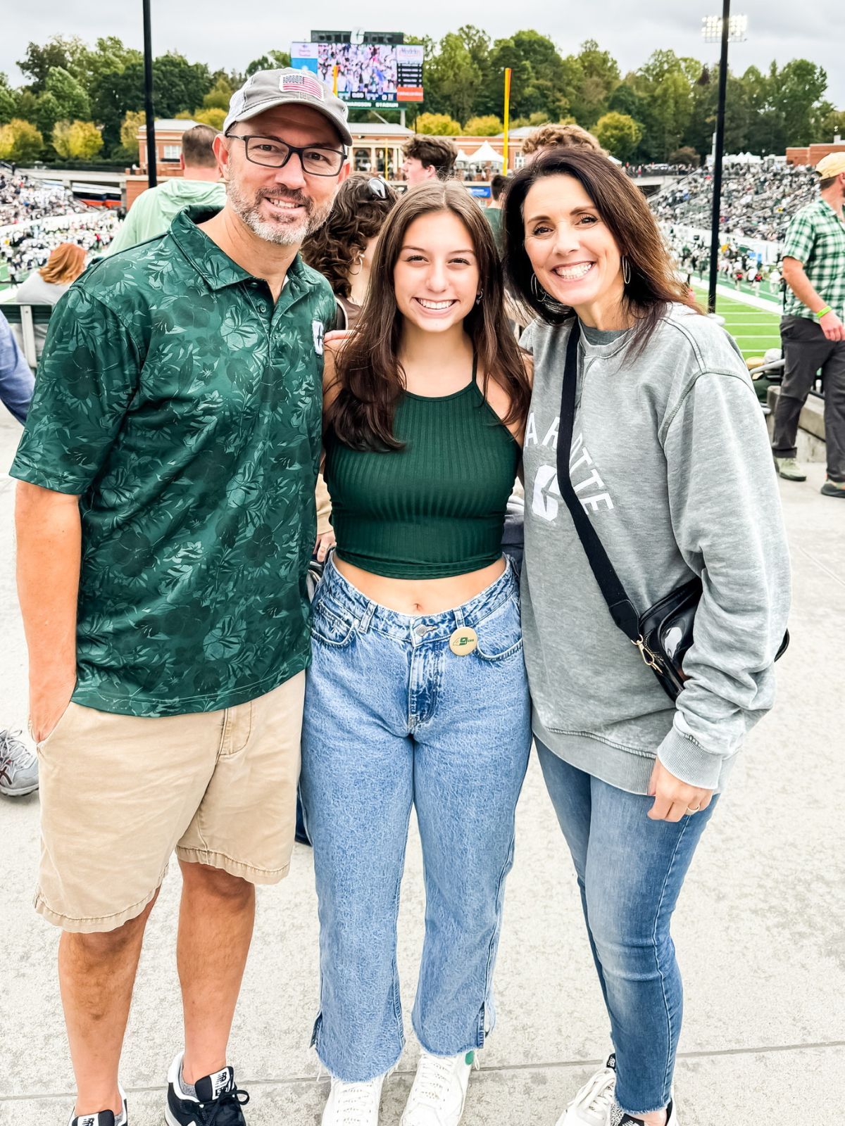 Three people stand together smiling at a stadium, with a football field and stands filled with people in the background. The group is casually dressed and appears to be enjoying a sporting event outdoors.