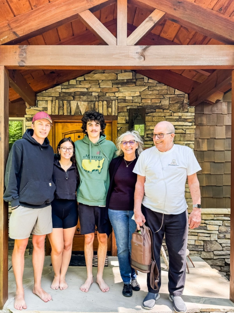 Five people stand smiling under a wooden porch in front of a stone house. Three young adults are on the left; two older adults, one holding a brown bag, are on the right. Everyone is dressed casually.