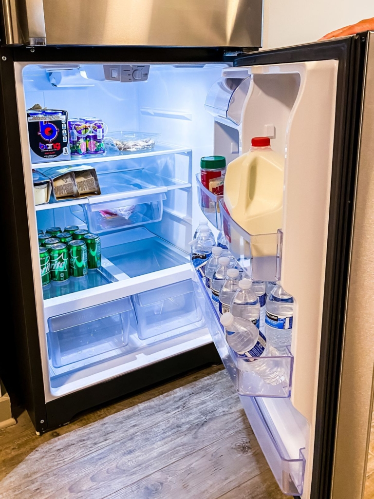 An open refrigerator with soda cans, bottled water, milk, condiments, and food containers on the shelves and door, illuminated by a bright light.